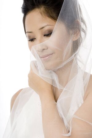 A young asian bride covers her face with her veil on white backgroundの写真素材