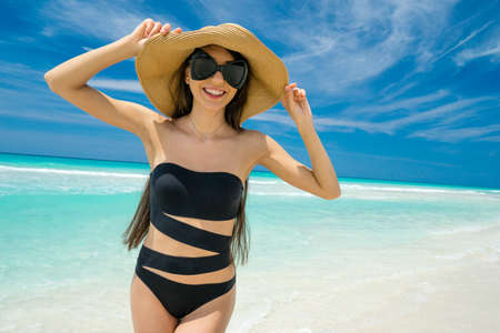Beautiful young girl in the straw hat, black swimsuit and sunglasses is smiling and standing on the beach on the background of azure caribbean sea. Hot summer day at the coastline of the Cuba.の写真素材