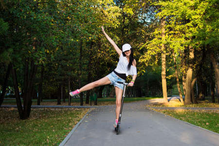 Close-up horizontal view athlete female rider in gray shoes with pink laces, shorts and white cap is riding on a black kick scooter on asphalt road in a park at summer sunny eveningの写真素材