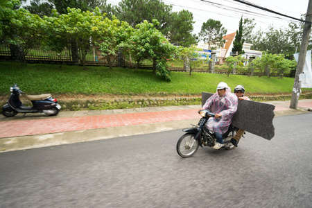DA LAT, VIETNAM - JUNE 23, 2016: Two asian men in raincoats ride the motorcycle on a street along park zone and bring metal plywoodのeditorial素材