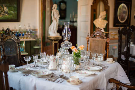 TRINIDAD, CUBA - APRIL 13, 2014: The served table with a white cloth, porcelain plates, china, crystal glasses and a vase. On a background there are marble statues and antique registration of the room at restaurantのeditorial素材