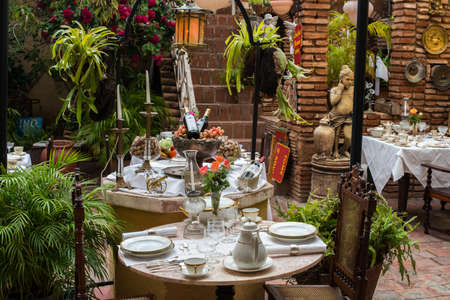 TRINIDAD, CUBA - APRIL 13, 2014: The served table with a white cloth, porcelain plates, china, crystal glasses and vase with flowers. There are marble statues and stone walls with a ladder in internal court yard on the streetのeditorial素材