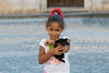HAVANA, CUBA - OCTOBER 03, 2016: Pretty latino little girl with red bow in curly hair is standing on a central square of the city and holding a black kitty in her arms at summer sunny dayのeditorial素材
