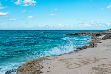 Beautiful view of rocky coast of azure coastline of Caribbean Sea in hot sunny summer day. Quiet small waves are splashing against coastal stones. Inscription Cuba on the sand. Clear serene blue skyの写真素材