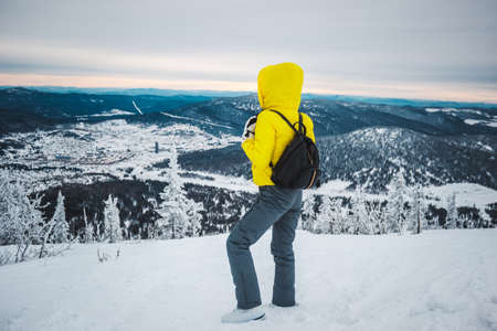 The young sport woman in a yellow jacket with hood and with black backpack stands a back sideways at top of the snow mountain and looks at the snow-covered valleyの写真素材