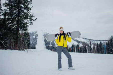 Beautiful caucasian young woman in yellow jacket and winter clothes smiles, stands and holds in hand snowboard on a background of ski resort and descent from the snow mountainの写真素材