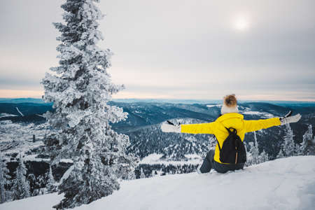 The young girl in a yellow jacket, white cap and with black backpack sits a back near fir-tree at top of the mountain, reaches for the sun and looks at the snow-covered valleyの写真素材
