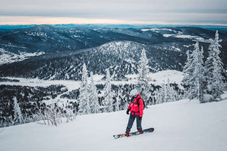SHEREGESH, RUSSIA - DECEMBER 06, 2016: Man snowboarder in red jacket and white helmet goes down from top of snow mountain on snowboard against the background of snow-covered valley and treesのeditorial素材