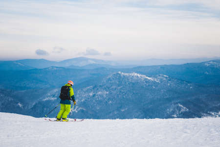 SHEREGESH, RUSSIA - DECEMBER 07, 2016: Man skier in bright green clothes with backpack goes down from top of snow mountain on skis against the background of snow-covered valley and amazing blue skyのeditorial素材