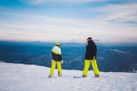 SHEREGESH, RUSSIA - DECEMBER 07, 2016: Two sportsmen snowboarders in bright yellow black costume stand and prepare for descent from snow top of the mountain on a snowboard down. Panoramic view of ski resort valley on a backgroundのeditorial素材
