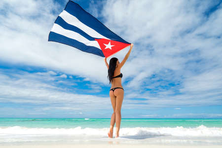 Beautiful young woman in black bikini swimsuit is standing a back on a beach on the background of azure sea and holding a flag of Cuba in her arms. Hot summer day at the caribbean coastline.の写真素材