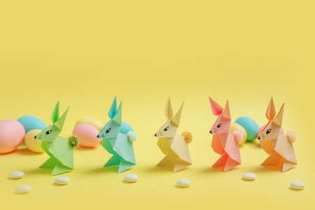 Paper Easter bunnies-origami made of colored paper and colored eggs on a delicate yellow background....