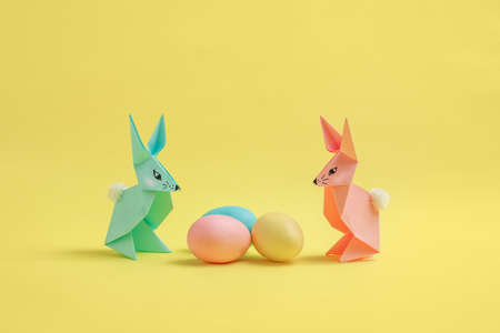 Paper Easter bunnies-origami made of colored paper and colored eggs on a delicate yellow background. The concept of the celebration of Easter, greeting card, crafts with your own hands.の写真素材