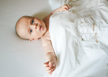 baby lies on a white sheet under a white blanketの写真素材