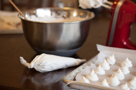 Homemade French meringue cookies/marshmallows on a rusty pan, pastry pouch, red mixer and chrome mixing bowl on brown wooden tableの写真素材