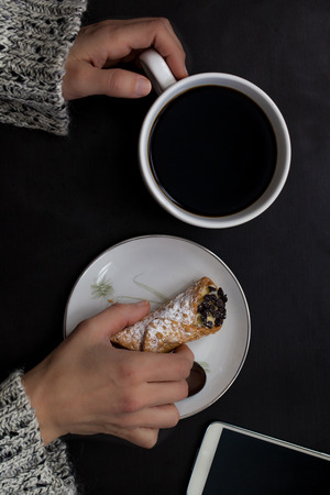 Caucasian woman hand holds coffee cup and smartphone with italian cannoli on plate on black backgroundの写真素材