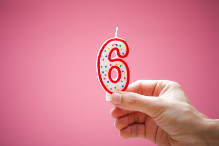 Hand holds colorful number 6 birthday cake candle on a pink background