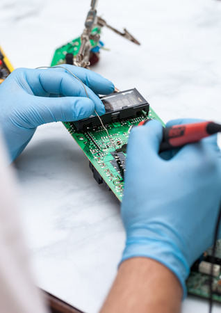 Hands wearing gloves solders and verifies computer circuit board using soldering iron and multimeterの写真素材