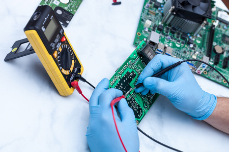 Hands wearing gloves solders and verifies computer circuit board using soldering iron and multimeterの写真素材