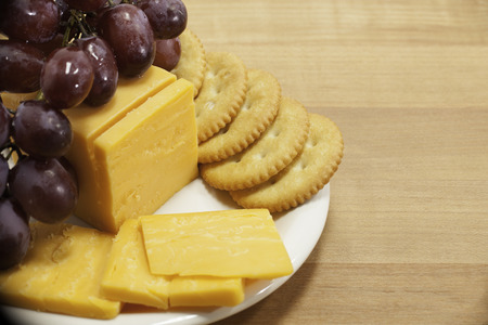 Cheddar cheese, round crackers, and red grapes are arranged on a small white plate with a...