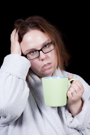 A woman with messy hair holds her head suggesting a headache   In her other hand is a coffee mug with hot tea   She wears glasses and a bathrobe の写真素材