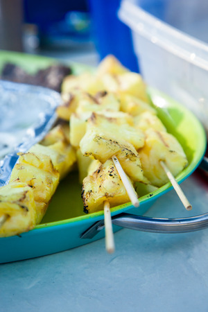 Several grilled pineapple kebabs sit on a green and blue tray at a barbecueの写真素材