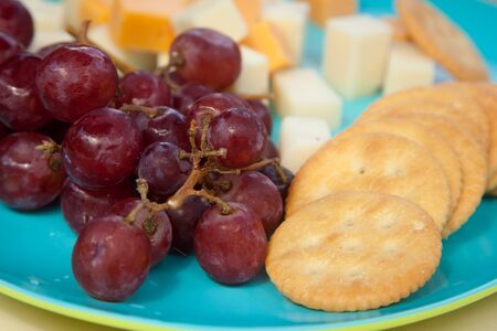 Fresh red grapes sit beside cubes of various cheeses and crackers on a blue and green plateの写真素材