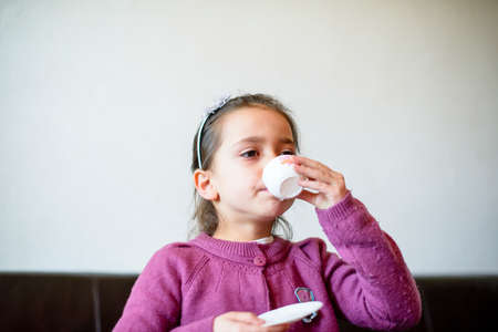 little girl plays, drinking tea on the sofa at home, lit by the windowの写真素材