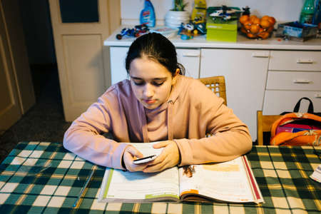 teenager does her homework in the home kitchen on the table helping herself with the smartphoneの写真素材