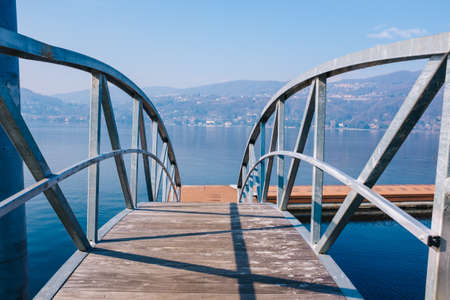 Pedestrian iron bridge with lake and mountains in a bright morning background, Ranco, Lombardy, Lake Maggiore, Italyの写真素材