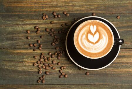 Top view of hot coffee Latte (or cappuccino) in a black cup with latte art and roasted coffee beans on the background of the wooden tableの写真素材