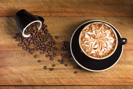 Top view of hot coffee Latte (or cappuccino) in a black cup with latte art and roasted coffee beans on the background of the wooden tableの写真素材