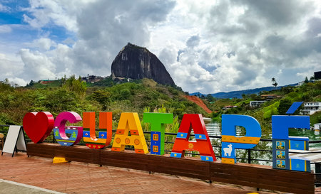 Guatape, Antioquia - Colombia. November 23, 2025. Colorful tourist sign with the imposing PeÃ±ol mountain in the background under a cloudy sky.のeditorial素材