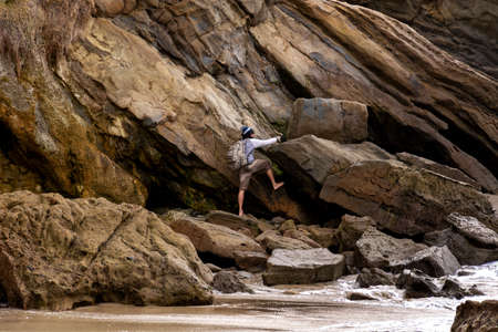 Laguna Beach, CA / USA - March 10, 2019: Man is climbing on the rocks of a cliff on the beach during low tide. The cove is surrounded by high cliffs with colorful striations in the rock.のeditorial素材