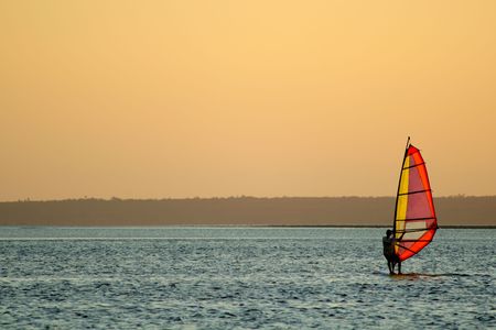 Backlit windsurfer at sunset on calm coastal water	の写真素材
