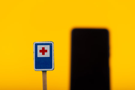 A toy wooden hospital warning sign is placed against a bright yellow background, with a smartphone softly out of focus, creating a conceptual and striking visualの写真素材