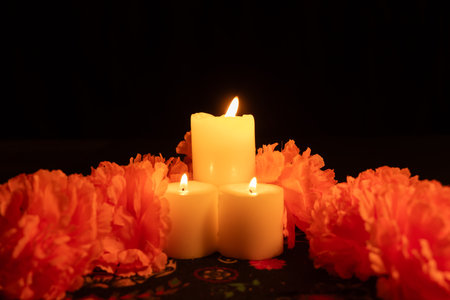 Vertical shot featuring three lit candles in the center, surrounded by vibrant orange marigolds. The scene, illuminated only by candlelight, rests on a DÃ­a de los Muertos themed tablecloth against a black background.の写真素材
