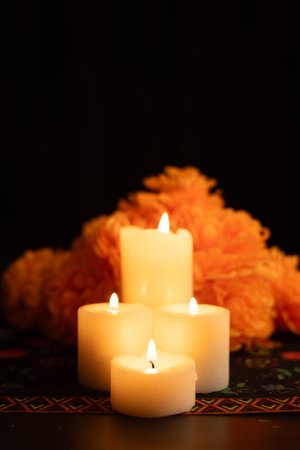 A vertical view of three lit candles encircled by vivid orange marigold flowers on a Day of the Dead themed tablecloth. The black background emphasizes the soft candlelight, creating a serene scene.の写真素材