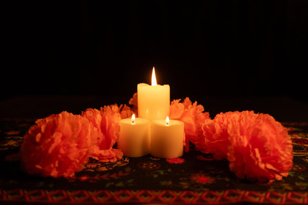 Three glowing candles are centered in a vertical image, surrounded by vibrant marigolds. The DÃ­a de los Muertos themed tablecloth and black background enhance the warm, intimate lighting of the scene.の写真素材
