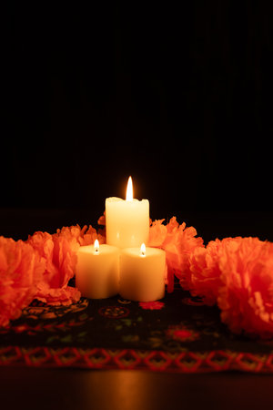 A vertical shot featuring three lit candles in the center of the image, surrounded by vibrant orange marigolds. The scene is illuminated by candlelight against a black background, set on a Day of the Dead-themed tablecloth.の写真素材