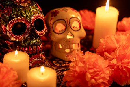 Two skulls on a Day of the Dead-themed tablecloth: one decorated with metallic flowers, the other hollow with a lit candle inside. Surrounded by orange marigolds and lit candles against a black background.の写真素材