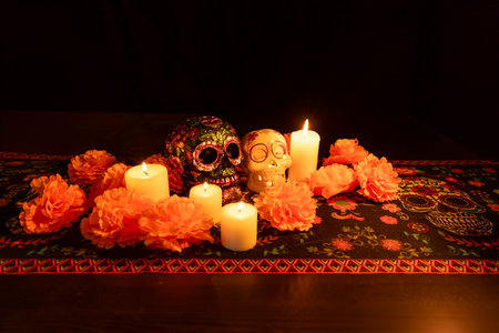 Featuring two skulls, one decorated with metal flowers and the other housing a lit candle. Surrounded by marigolds and candles, all presented on a Day of the Dead themed tablecloth with a dark background.の写真素材