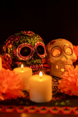 Vertical composition featuring two skulls on a Day of the Dead tablecloth: one decorated with metallic flowers, the other hollow with a lit candle. Marigold flowers and additional candles create a festive atmosphere against a black background.の写真素材