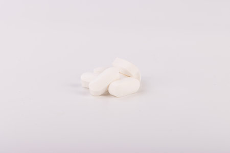 Close-up of white pills scattered on a plain white background. The focus is on the pills, emphasizing their texture and shape, ideal for medical and health-themed contentの写真素材
