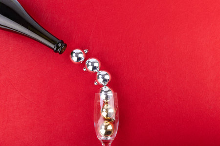 A bottle gracefully pours into a glass filled with shimmering silver ornaments, mimicking bubbly foam, set against a rich red backdropの写真素材