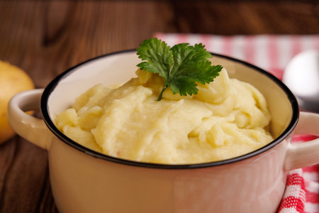 Mashed potatoes in a bowl, accompanied by whole potatoes, a spoon, and a red napkin on a brown wooden tableの写真素材