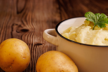A bowl of freshly made mashed potatoes next to whole raw potatoes on a brown wooden table. The warm, creamy texture contrasts with the rustic backgroundの写真素材