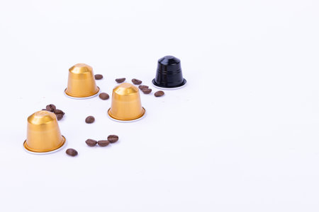 A collection of elegant golden and black coffee capsules rests beside scattered coffee beans on a pristine white surfaceの写真素材