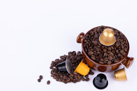 Brown bowl with coffee beans and a capsule on top, surrounded by scattered beans and colorful capsules on a plain white backgroundの写真素材
