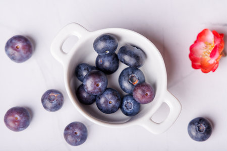 Scattered blueberries adorn a white marble table, highlighting a porcelain bowl brimming with fresh berries, offering a vibrant and delicious sightの写真素材